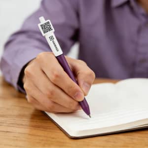 A purple click pen featuring a QR code, held by a person writing in a notebook on a wooden surface.