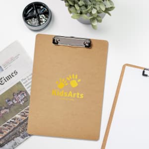 A wooden clipboard in a light brown colour with a silver metal clip. It features a logo and is placed on a desk with greenery.