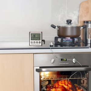 A digital oven thermometer in silver with a probe, placed on a kitchen countertop beside a pot and oven.