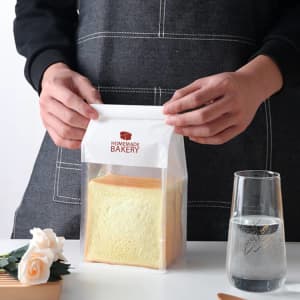 A transparent sealing storage bag holding a loaf of bread, featuring a logo and a white top closure.