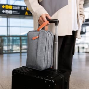 A grey classic toiletry bag with a tan handle sits atop a black suitcase, at an airport terminal.