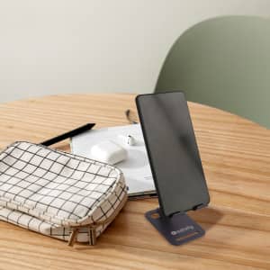 A foldable metal mobile phone stand in black, with a smartphone displayed, on a wooden table along with a bag and accessories.