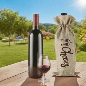 A wine drawstring bag in natural fabric beside a red wine bottle and a glass of wine outdoors in a garden setting.