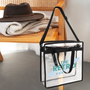 A transparent tote bag with black straps sits on a wooden chair next to a hat and a folded sweater.