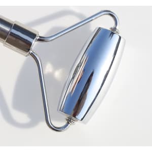A stainless steel facial roller with a shiny, reflective surface and a curved design. It has a logo on the handle.