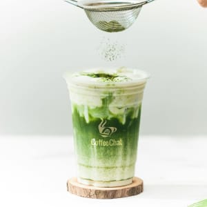 A clear plastic cup filled with a green drink and topped with frothy cream, featuring a logo on the cup.