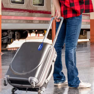 A person holds a black suitcase with a blue coloured silicone luggage tag attached, standing at a train station.