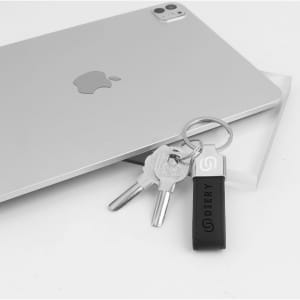 A metal keyring in black PU leather with two silver keys, placed beside a silver tablet. It features a logo.