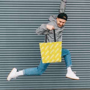 A person jumps while holding a vibrant yellow tote bag featuring a logo, set against a striped grey background.