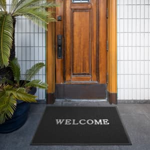 A black floor mat with a logo in front of a wooden door, surrounded by greenery.