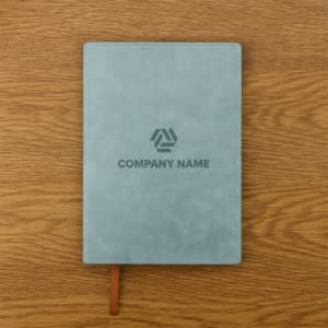 A soft PU leather notebook in a muted green colour with a logo on the cover, placed on a wooden surface.