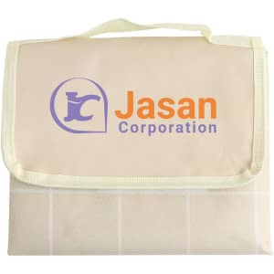 A cream picnic blanket with a green trim and a logo on the front, folded and secured with a handle.