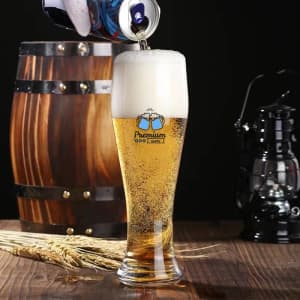 A tall, clear glass filled with pale beer and a frothy head, displayed alongside a wooden barrel and a lantern.