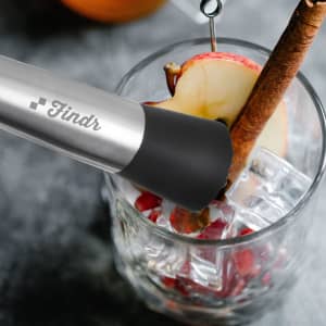 A stainless steel cocktail muddler with a black silicone tip, alongside a glass filled with ice, apple slices, and a cinnamon stick.