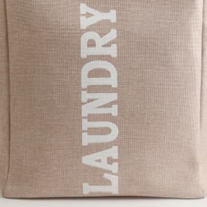 A laundry basket in a beige fabric with white lettering, featuring a sturdy handle and a simple design.