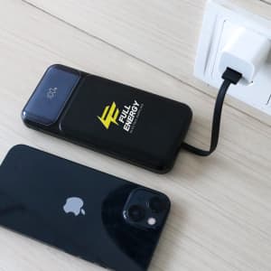 A black power bank with a logo is charging near a phone, connected to a wall socket with a cable.