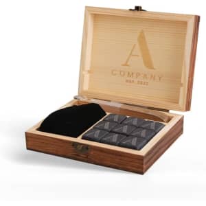 A wooden box containing grey whiskey stones, a pair of tongs, and a black storage pouch, featuring a logo on the lid.
