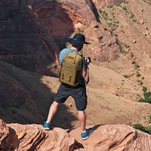 A man wearing a black hat and blue shirt stands on rocks, with a tan waxed canvas camera sling bag on his back.