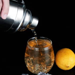 A drink bottle in metallic finish pouring a cocktail into a glass with ice, accompanied by a lemon on the side.
