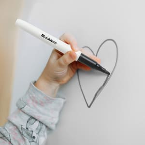 A hand holding a black marker, drawing a heart shape on a light surface. The marker has a white body and a logo.