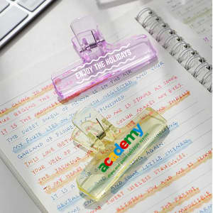 Translucent plastic clips in purple and yellow resting on handwritten notes, with a notebook and a computer keyboard in the background.