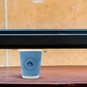 A double wall paper cup in blue, featuring a logo, sits on a wooden table against a textured background.