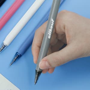 A hand holding a grey plastic pen with a transparent barrel, surrounded by pink, white, blue, and purple pens, and a notebook.