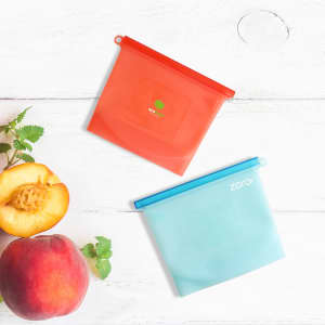 Two silicone reusable storage pouches in red and blue, placed on a wooden surface alongside a peach and a sprig of herbs.