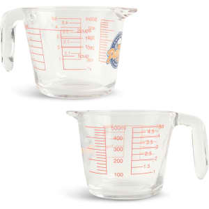 A clear glass baking measuring jug with red measurement markings, featuring a handle and a logo.