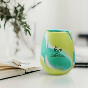 A silicone cup in green and blue marbled design, featuring a logo, on a table with a notebook and a plant.