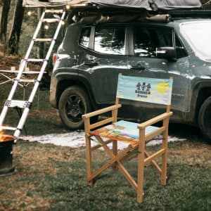 A folding director's chair with a light blue seat featuring a beach design, set near a vehicle in a forested area.