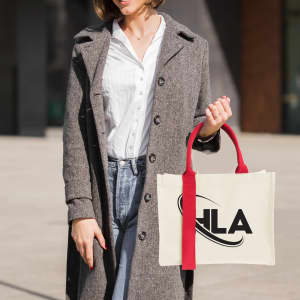 A woman holds a cream-coloured horizontal canvas tote bag with red handles, featuring a logo.