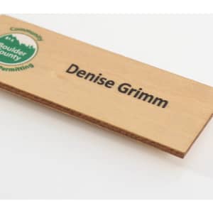 A wooden name badge in a natural finish featuring a logo and black text.