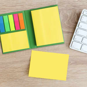 A compact sticky notepad with yellow pages and colourful sticky flags, placed on a wooden surface beside a keyboard.
