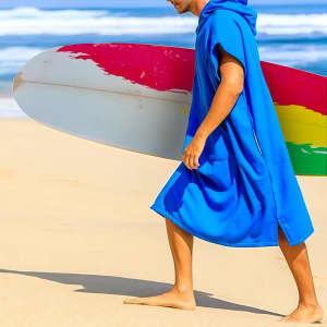 A blue cotton poncho-style towel with a hood, being worn by a person holding a surfboard on the beach.