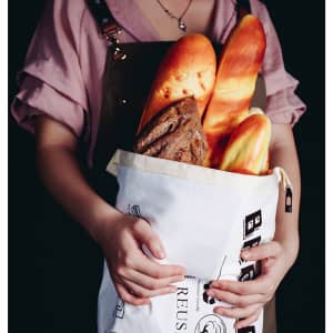 A linen bread bag in white, filled with various types of bread, held by a person in a pink shirt.