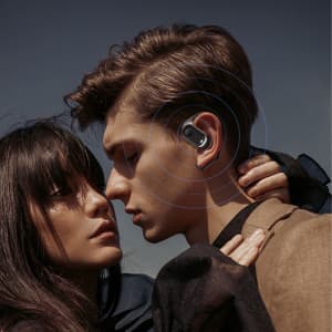 Open-ear headphones in silver are worn by a person, who is facing another person, against a clear sky backdrop.