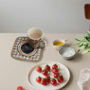 A medium quilted pot holder in a checkered pattern with a logo, placed on a table with a mug and fresh tomatoes.