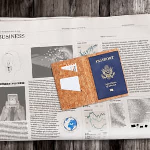 A cork passport holder with a blue passport inside, resting on a newspaper background. It features a logo on the inside.