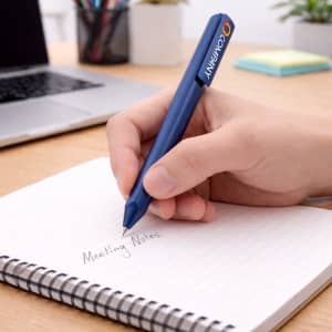 A blue triangular grip pen is held above a notepad with handwritten notes on a wooden desk with a laptop in the background.