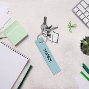 A light blue woven key tag attached to keys, surrounded by stationery and a small plant.