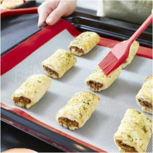 A silicone baking mat in red is placed on a baking tray, with savoury pastries being brushed with a red tool.