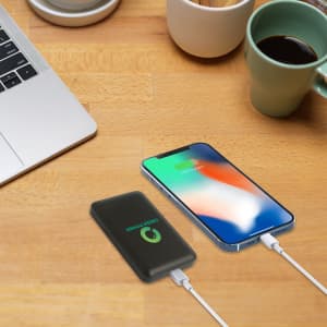 A black 10000mAh power bank with a logo beside a smartphone, on a wooden desk with a coffee cup.
