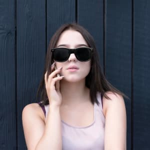A pair of black classic sunglasses displayed against a dark wood background, worn by a young woman.