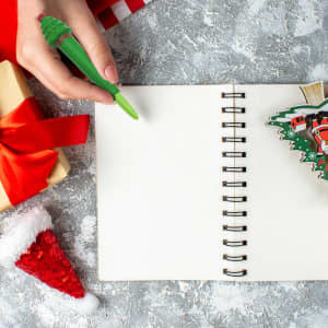 A green tree-shaped stress reliever pen held above a blank spiral notebook, surrounded by a gift and festive decorations.