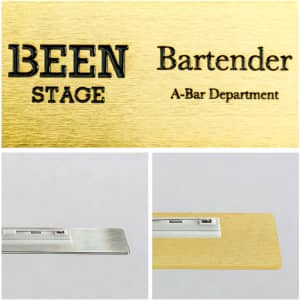 A metal name badge in gold with a brushed finish, featuring a silver backing. Includes a logo.