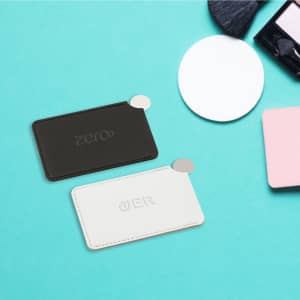 A black and white stainless steel portable mirror with a sleek design, placed on a light blue surface alongside beauty tools.