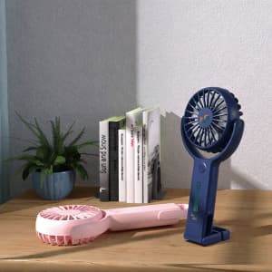 A handheld fan in pink and navy blue with a 180° adjustable angle, sitting on a wooden table beside books and a plant.