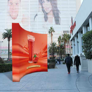 A curved fabric popup display in vibrant orange featuring a beverage design, set outdoors near a building.