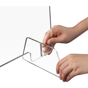 A clear acrylic personal protection screen is being assembled, held by hands at the base.